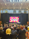 A photograph of the attendees gathering and chatting at the beginning of the BookUp conference. They are seated in many rows, and there is a screen showing the BookUp logo above the stage at the front of the room