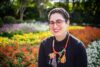 An author photograph of Anna Jacobson, who is pictured in front of a garden with a range of flowering plants